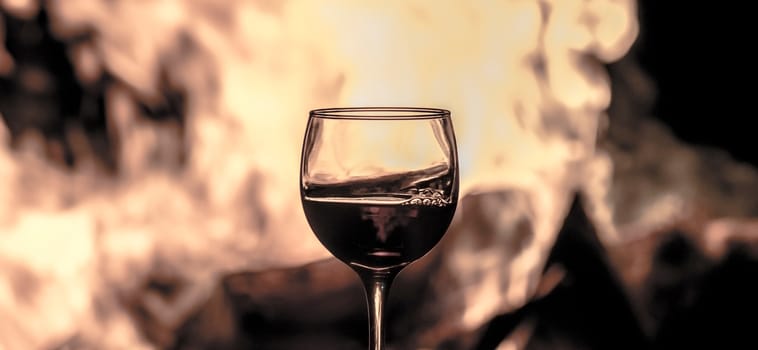 A glass of red wine in front of a camp fire, allowing see through it all the color shades of the flames