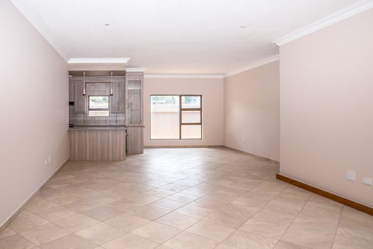 A vacant livingroom of a newly build house, with tiled floors, newly painted walls, and part of the kitchen in the background.