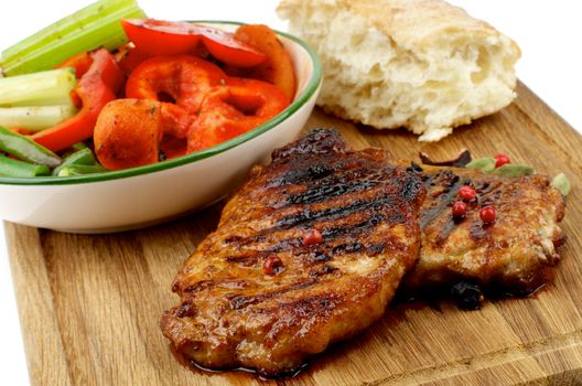 Delicious Roasted Pork Steaks with Vegetable Stocks and Flap of Bread on Wooden Cutting Board isolated on white background. Focus on Steaks