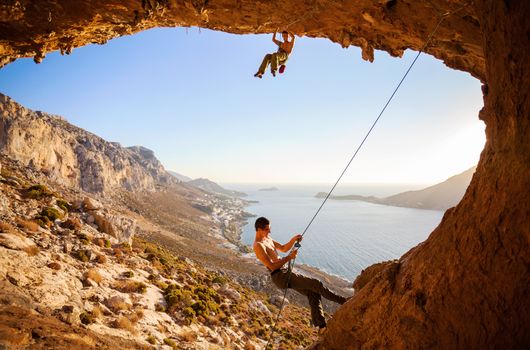 Male rock climber climbing on a roof in a cave, his partner belaying