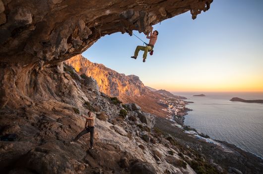 Male rock climber climbing on a roof in a cave, his partner belaying