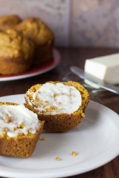 A cut and buttered pumpkin muffin opn a white plate with a plate of muffins in the background.