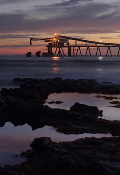 The gravel loader at Bass Point at dawn.   A 400m long pier.  Cargo ships dock alongside for filling  Mostly silhouetted against the dawn sky except where its lights illuminate it. Long exposure,