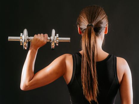 Photo of the back of a young woman doing a shoulder press with a dumbbell over a dark background.