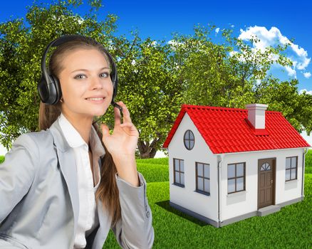 Businesswoman in headset, her hand on microphone, looking at camera, smiling. Small house in green landscape as backdrop