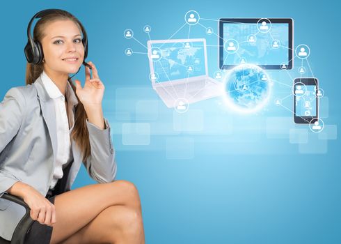 Businesswoman in headset, sitting on office chair, her hand on microphone, looking at camera, smiling. Globe, Network with people icons, laptop, tablet PC and smartphone beside, on light blue background