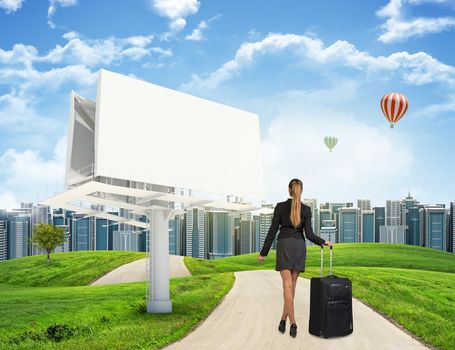 Businesswoman with wheeled suitcase takes a step forward on the road among green hills, against distant cityscape and balloons in the sky. Blank billboard beside. Looking at camera. Business concept