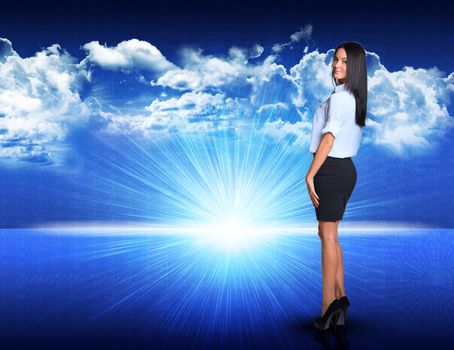 Businesswoman standing against digitally generated spacy blue landscape with rising sun and cloudy sky, looking back over her shoulder