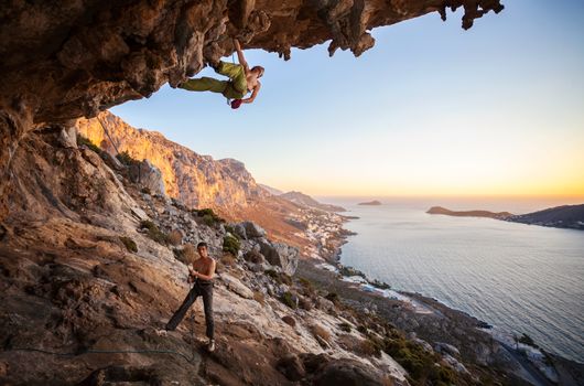 Male rock climber climbing on a roof in a cave, his partner belaying