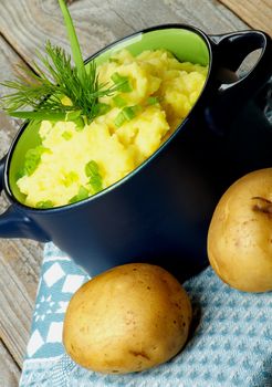 Creamy Homemade Mashed Potato with Dill and Spring Onion in Casserole on Blue Napkin with Raw Potatoes closeup on Rustic Wooden background