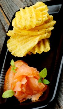 Snack with Delicious Smoked Salmon and Rifled Potato Chips closeup on Black Plate on Rustic Wooden background