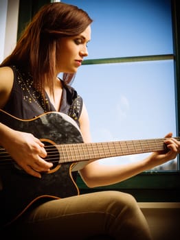 Photo of a beautiful brunette female playing an acoustic guitar by a window.
