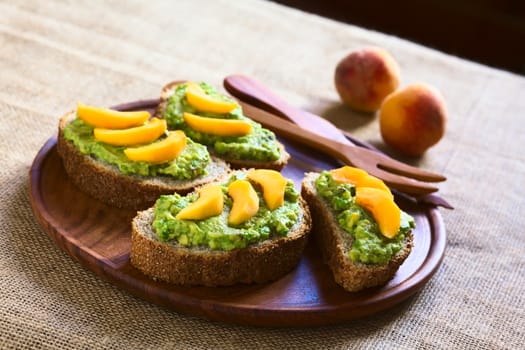 Wholegrain bread slices with avocado spread and peach slices served on wooden plate photographed with natural light (Selective Focus, Focus in the middle of the bread in the middle of the image)