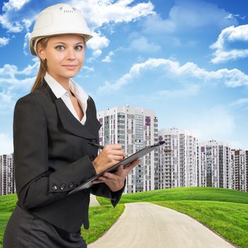 Businesswoman standing on the road among green hills, wearing hard hat, writing on clipboard, looking at camera. High-storey buildings, sky and clouds as backdrop