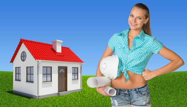 Woman holding hard hat and drawing rolls, looking at camera, smiling. Small house on green field and blue sky as backdrop