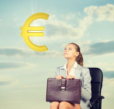 Businesswoman sitting on office chair, holding briefcase on her knees, looking at golden euro sign in the air, against blue sky with clouds