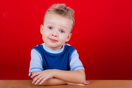 Small boy in blue T-shirt, sitting at the table. On a red background