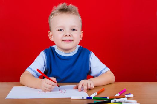Small boy draws at the table. On a red background