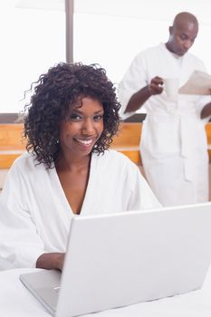 Pretty woman in bathrobe using laptop at table with partner in background at home in the kitchen