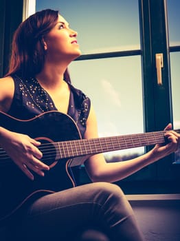 Photo of a beautiful brunette female playing an acoustic guitar by the window. Heavily filtered.

