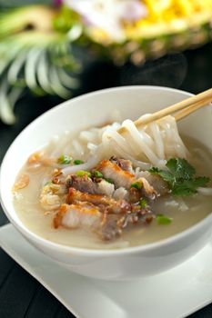 Closeup of a person eating Thai style crispy pork rice noodle soup from a bowl with chopsticks. Pineapple fried rice in the background.