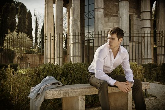 Handsome young man in European city, sitting on stone bench, looking away to a side