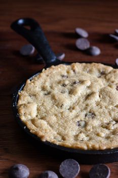 A personal-sized chocolate chip cookie in a cast iron pan with chocolate chips in the background.