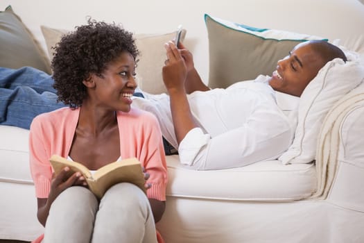 Happy couple relaxing together reading book and using smartphone at home in the living room