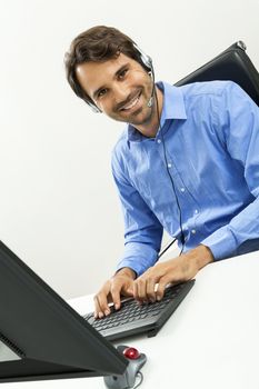 Attractive unshaven young man wearing a headset offering online chat and support on a client services of help desk as he types in information on his computer