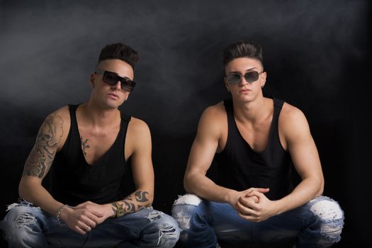 Two hip trendy male friends in studio shot sitting on the floor, wearing sunglasses and black tanktops