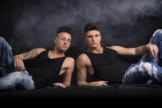 Two hip trendy male friends in studio shot laying on the floor, wearing black tanktops