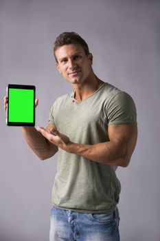 Smiling young man holding and showing tablet PC or ebook reader, standing on grey background