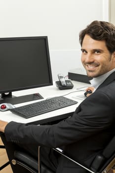 Stylish businessman in a suit sitting at his desk in the office chatting on the phone with a view of his blank computer monitor