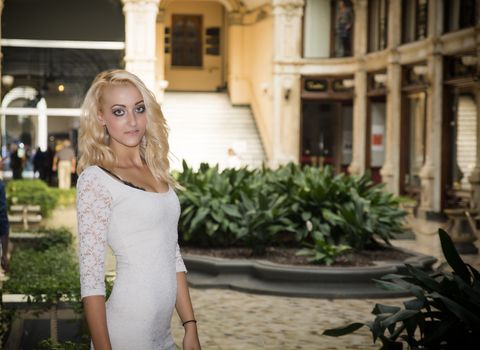 Elegant pretty blonde young woman standing in white dress in posh city setting in Europe