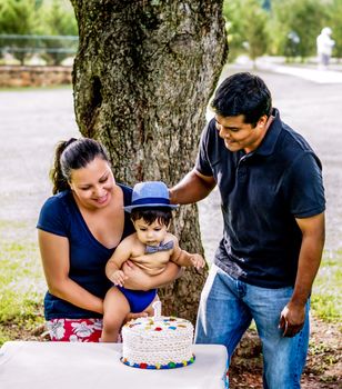 Latino mother, father, and baby outside by a tree with a birthday cake