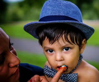 Latino baby wearing a blue hat and bow tie eating frosting from his mother's finger