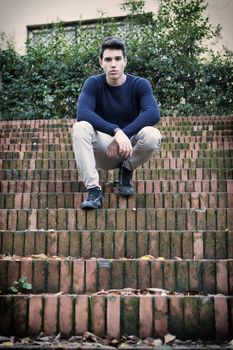Handsome young man sitting outdoor on old brick stairs looking confident at camera
