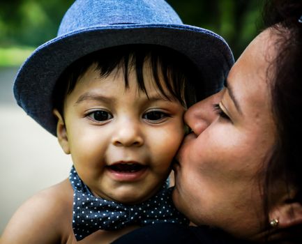 Smiling latino baby wearing a blue hat and bow tie being kissed on cheek by female adult family member