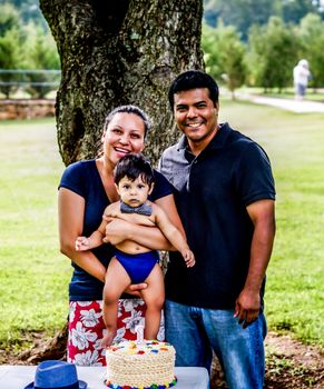 Latino mother, father, and baby with frosting on his face outside by a tree with a birthday cake