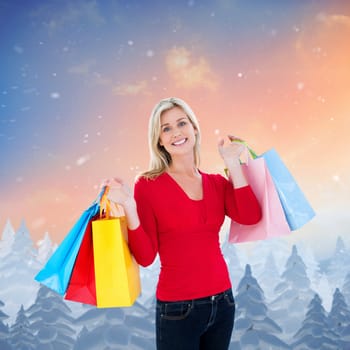 Happy blonde holding shopping bags against snow falling on fir tree forest
