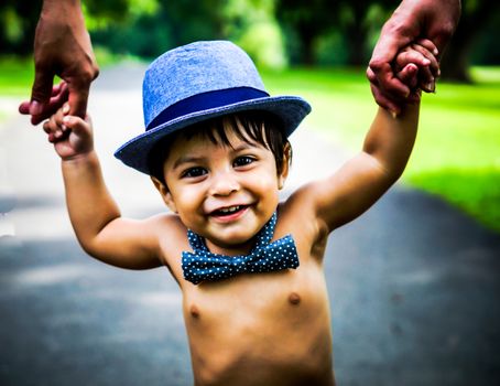 Smiling latino baby wearing a blue hat and bow tie walking outside and holding hands with adults