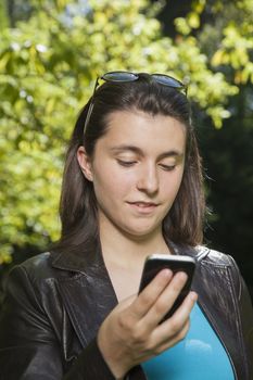pregnant young woman with blue shirt and brown leather jacket calling phone over green trees background street