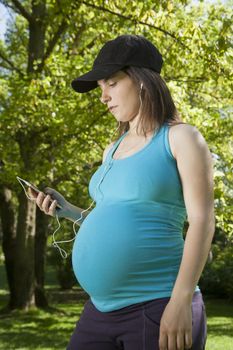 pregnant young woman blue shirt listen to phone or music player with white earphones over green tree background