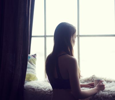 Woman in the morning. woman with neat body is holding a cup with hot tea or coffee and looking at the sunrise standing near the window in her home and having a perfect cozy morning