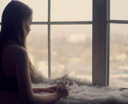 Woman in the morning. woman with neat body is holding a cup with hot tea or coffee and looking at the sunrise standing near the window in her home and having a perfect cozy morning