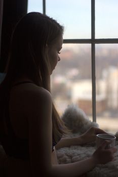 Woman in the morning. woman with neat body is holding a cup with hot tea or coffee and looking at the sunrise standing near the window in her home and having a perfect cozy morning