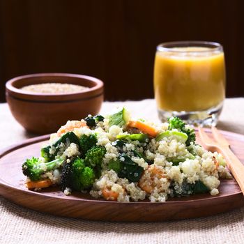 Cooked white quinoa seeds with fried vegetables (carrot, broccoli, spinach, zucchini, corn) on wooden plate, sesame seeds and mango juice in the back, photographed with natural light (Selective Focus, Focus one third into the dish)