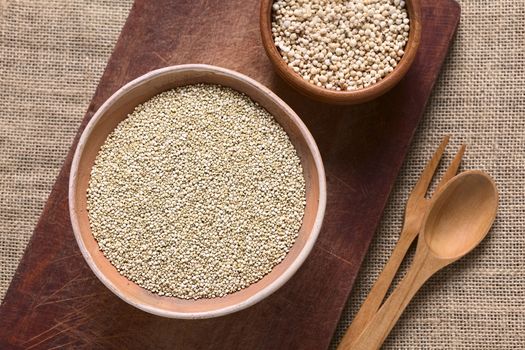 Overhead shot of raw white quinoa (lat. Chenopodium quinoa) grain seeds in bowl with popped quinoa cereal on wooden board photographed with natural light