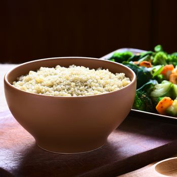 Cooked white quinoa seeds in bowl with cooked vegetables in the back photographed with natural light (Selective Focus, Focus one third into the quinoa)