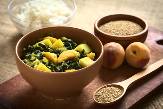 Bowl of spinach, peach and potato curry dish with sesame seeds and cooked rice in the back photographed with natural light (Selective Focus, Focus one third into the curry dish)